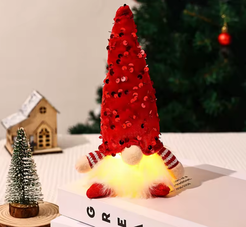 Decorative gnome with a red hat and striped scarf, sitting on a box with a small Christmas tree and house in the background.