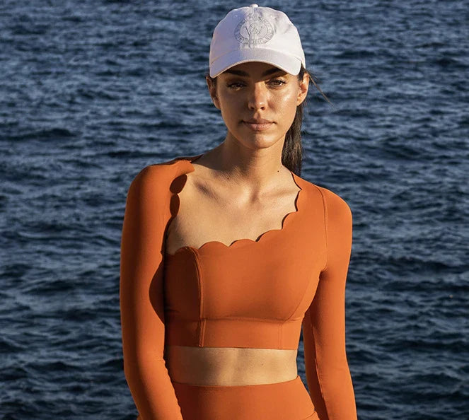 Woman in an orange outfit and white cap standing in front of a body of water