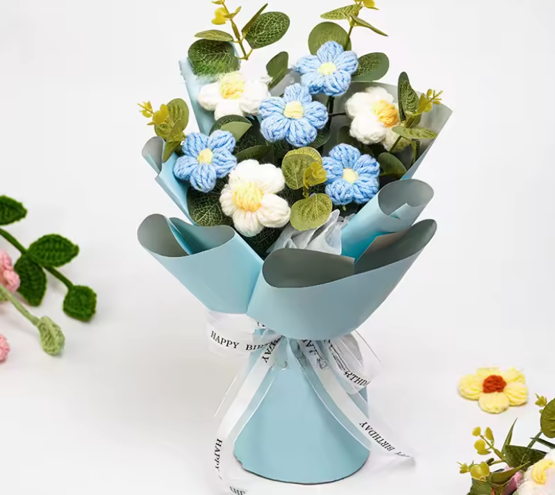 Bouquet of knitted flowers in blue, white, and yellow with a light blue cone-shaped holder on a white background.